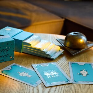 Może przedstawiać: Turkusowa talia kart tarota z napisem "Merry Tarot deck" na pudełku. Wyświetlanych jest kilka kart tarota, z fantazyjnymi ilustracjami. Złoty przedmiot dekoracyjny spoczywa na małej kwadratowej miseczce.