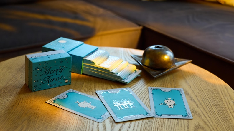Może przedstawiać: Turkusowa talia kart tarota z napisem "Merry Tarot deck" na pudełku. Wyświetlanych jest kilka kart tarota, z fantazyjnymi ilustracjami. Złoty przedmiot dekoracyjny spoczywa na małej kwadratowej miseczce.