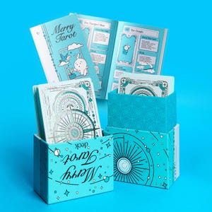 Może przedstawiać: Turkusowy zestaw kart Merry Tarot z pasującym przewodnikiem. Pudełko na talię zawiera napis "Merry Tarot deck" w fantazyjnej czcionce. Przewodnik i karty zawierają ilustracje kr&oacute;lika i inne fantazyjne wzory. Tło jest jednolicie niebieskie.