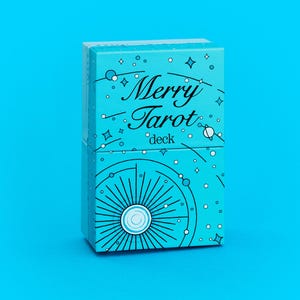 Może przedstawiać: Turkusowe pudełko na talię kart tarota z napisem "Merry Tarot deck" w ozdobnej czcionce. Pudełko jest ozdobione białymi gwiazdami, kropkami i niebiańskim wzorem. Tło jest w jednolitym turkusowym kolorze.