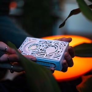 May include: A white rectangular deck of cards box featuring intricate laser-cut patterns and the word GARDEN printed on the side. The box has a layered design with visible cutouts revealing colourful details underneath the white surface.