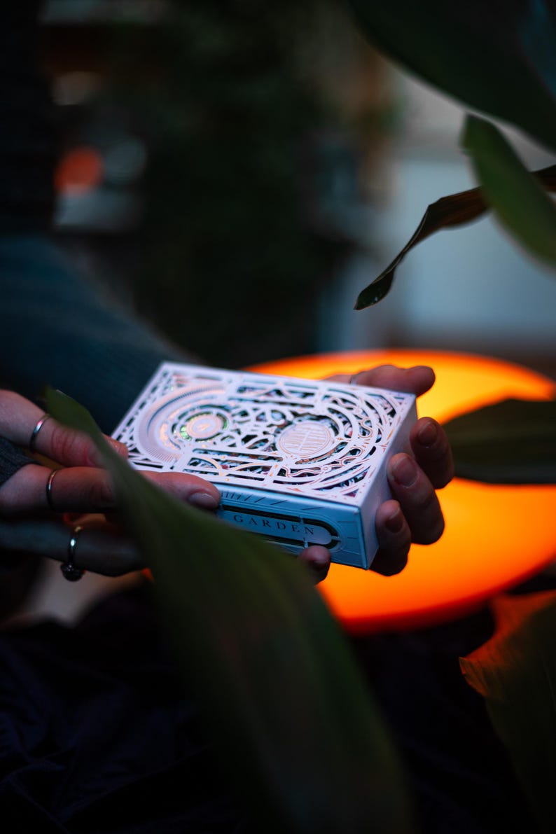 May include: A white rectangular deck of cards box featuring intricate laser-cut patterns and the word GARDEN printed on the side. The box has a layered design with visible cutouts revealing colourful details underneath the white surface.