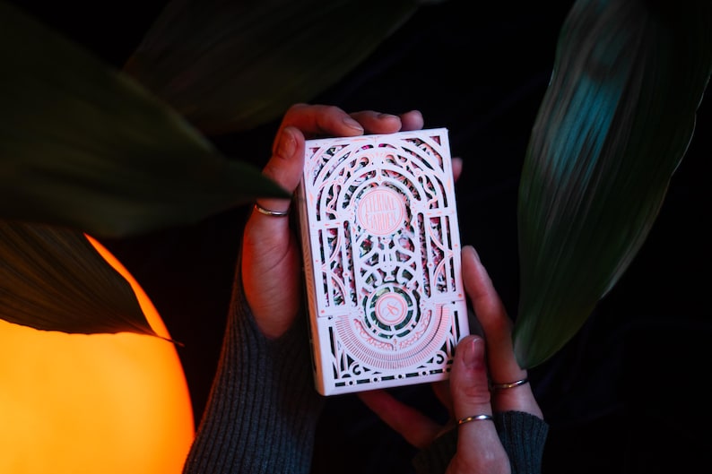 May include: A rectangular deck of playing cards featuring an intricate white laser-cut paper sleeve with arched architectural patterns and delicate pink accents. The design includes a circular emblem at the centre and geometric filigree details.