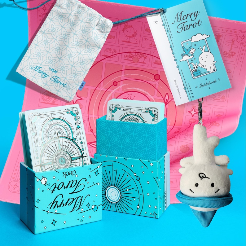 Może przedstawiać: Kompletny zestaw kart Merry Tarot, zawierający przewodnik, woreczek ze sznurkiem i pluszowy urok. Zestaw zawiera r&oacute;żową matę z okrągłym wzorem i jasnoniebieskie pudełko z napisem "Merry Tarot deck".