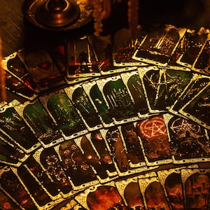 May include: A deck of tarot cards fanned out, featuring illustrations with gold accents. The cards have a dark, gothic aesthetic with images of forests, figures, and symbols. The overall color scheme is dark with green and gold.
