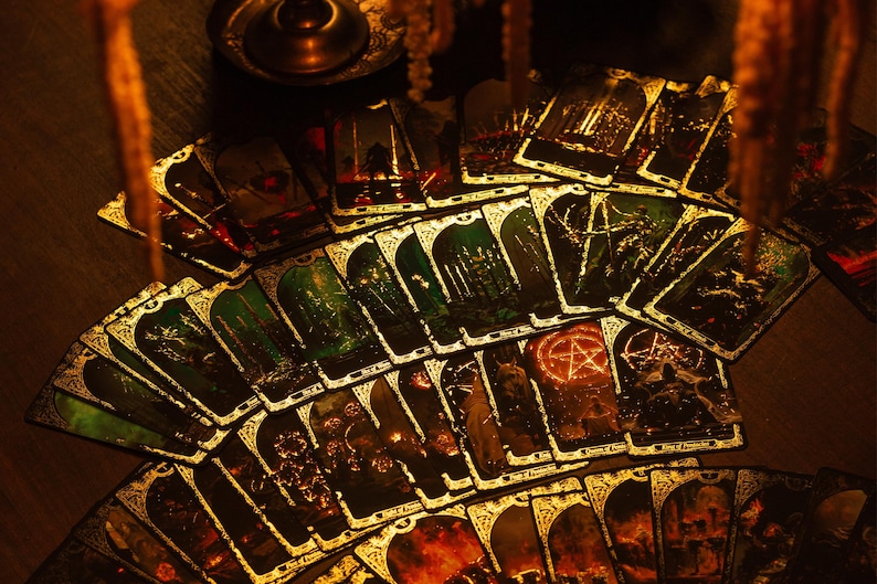May include: A deck of tarot cards fanned out, featuring illustrations with gold accents. The cards have a dark, gothic aesthetic with images of forests, figures, and symbols. The overall color scheme is dark with green and gold.