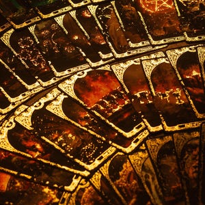 May include: A fan of tarot cards with gold trim and intricate illustrations. The cards have a dark, mystical theme with images of fire, figures, and celestial symbols. The overall color palette is warm, with shades of brown, gold, and red.