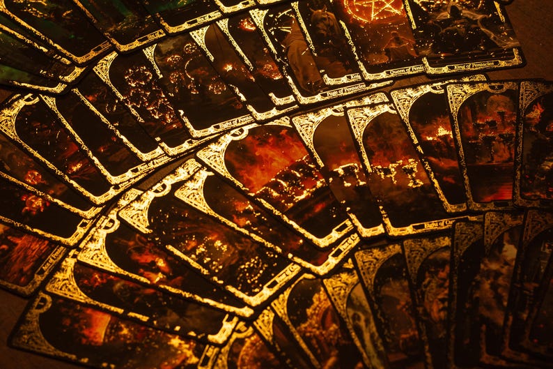May include: A fan of tarot cards with gold trim and intricate illustrations. The cards have a dark, mystical theme with images of fire, figures, and celestial symbols. The overall color palette is warm, with shades of brown, gold, and red.