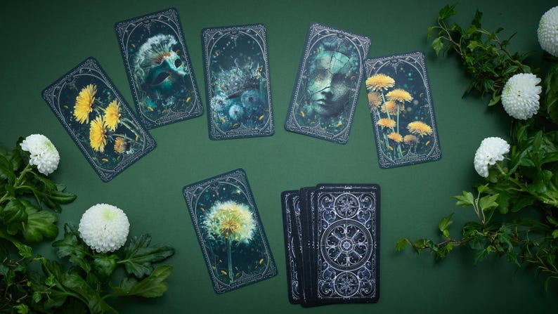 May include: A set of rectangular tarot cards featuring dark, moody illustrations of yellow dandelions, a human face, and celestial patterns. The cards are arranged in a spread with one deck stacked, surrounded by white chrysanthemum flowers and green leaves.