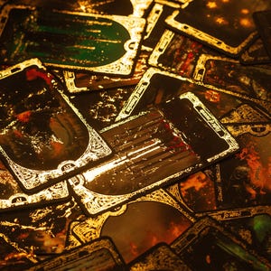 May include: A collection of tarot cards with gold trim and dark, detailed illustrations. The cards are arranged in a pile, with some showing images of fire and other mystical scenes. The overall color scheme is dark with gold accents.