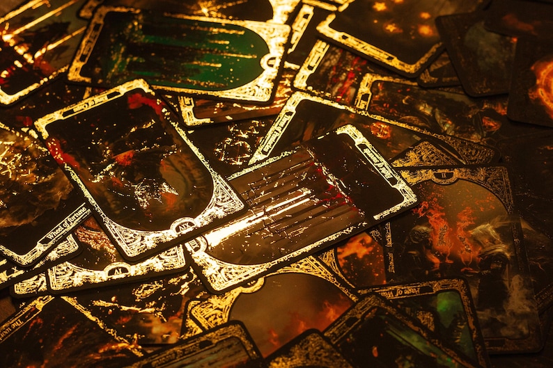 May include: A collection of tarot cards with gold trim and dark, detailed illustrations. The cards are arranged in a pile, with some showing images of fire and other mystical scenes. The overall color scheme is dark with gold accents.