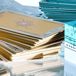 May include: A stack of tarot cards with gold and cream-colored designs, featuring celestial illustrations. The cards are arranged near a light blue box with the word "Merry" and other decorative elements. Other stacks of cards are visible.