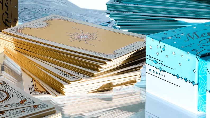 May include: A stack of tarot cards with gold and cream-colored designs, featuring celestial illustrations. The cards are arranged near a light blue box with the word "Merry" and other decorative elements. Other stacks of cards are visible.