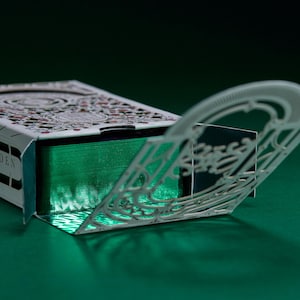 May include: A deck of playing cards housed in a white box with intricate laser-cut filigree patterns. The box is partially open, revealing a stack of cards with metallic green edges. The design features detailed floral motifs and geometric lines.