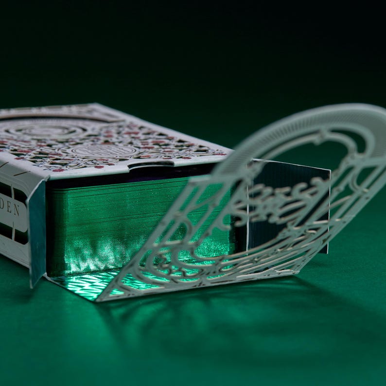 May include: A deck of playing cards housed in a white box with intricate laser-cut filigree patterns. The box is partially open, revealing a stack of cards with metallic green edges. The design features detailed floral motifs and geometric lines.