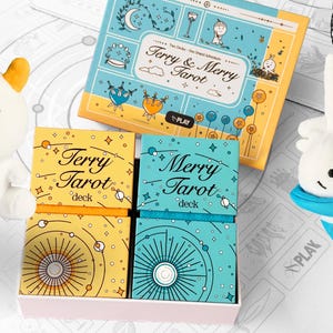 Może przedstawiać: Zestaw kart tarota z pasującą pluszową zabawką. Zestaw zawiera dwie talie z napisami "Terry Tarot" i "Merry Tarot", przewodnik i dwie pluszowe zabawki. Talie znajdują się w r&oacute;żowym pudełku. Pluszowe zabawki są białe z ż&oacute;łtymi i niebieskimi akcentami.