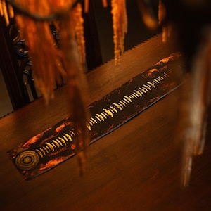 May include: A long, rectangular wooden object with a decorative design. The design features a series of gold-colored, tooth-like shapes along the center, with a circular pattern at one end. The object is set on a wooden surface.