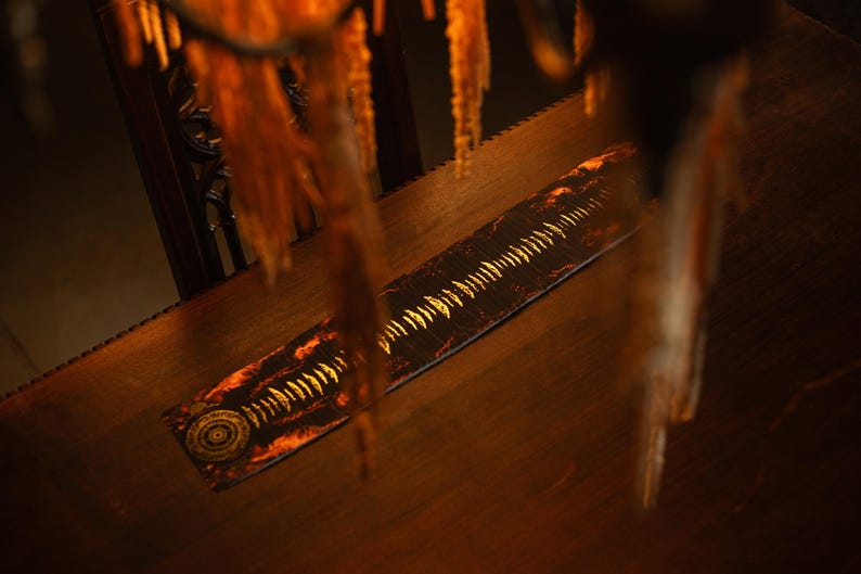 May include: A long, rectangular wooden object with a decorative design. The design features a series of gold-colored, tooth-like shapes along the center, with a circular pattern at one end. The object is set on a wooden surface.