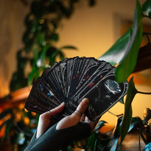 May include: A fan of black tarot cards featuring intricate white line art borders and detailed botanical illustrations of flowers. The cards are held in a spread, showcasing the dark aesthetic and the ornate, decorative framing on each individual card.