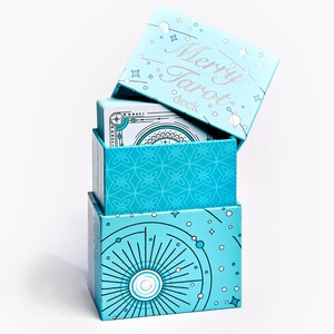 Może przedstawiać: Stos trzech jasnoniebieskich pudełek do kart tarota. Na g&oacute;rnym pudełku widnieje napis "Merry Tarot deck" srebrnym pismem. Pudełka zdobione są niebiańskimi i geometrycznymi wzorami w kolorze białym i turkusowym. G&oacute;rne pudełko jest lekko otwarte.