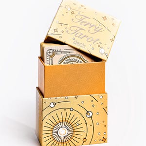 May include: Stacked tarot card boxes in shades of gold and orange. The top box is labeled "Terry Tarot deck" in silver lettering. The boxes feature celestial and geometric designs, with a white background.