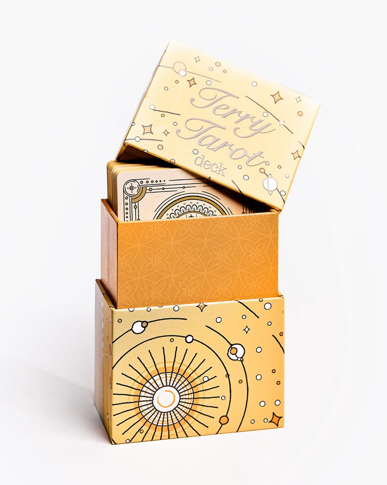 May include: Stacked tarot card boxes in shades of gold and orange. The top box is labeled "Terry Tarot deck" in silver lettering. The boxes feature celestial and geometric designs, with a white background.
