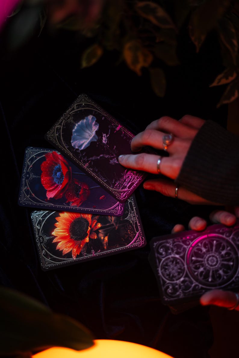 May include: A set of rectangular tarot cards featuring detailed floral illustrations including a bright yellow sunflower, a vibrant red poppy, and a pale blue morning glory, all framed with ornate silver borders on a dark background.