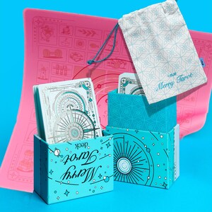Może przedstawiać: Turkusowy zestaw kart "Merry Tarot" z r&oacute;żową matą i wzorzystym workiem ze sznurkiem. Pudełka na talię mają wz&oacute;r promieni słonecznych, a karty mają wz&oacute;r r&oacute;ży wiatr&oacute;w. Torba i mata mają jasnoniebieską i r&oacute;żową kolorystykę.