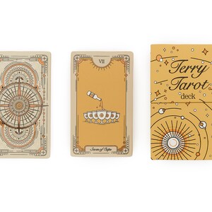 May include: A tarot card deck with three visible elements. Two cards and a box are displayed. The cards have beige and orange designs. The box is gold with the text "Terry Tarot deck". The cards have intricate line art.