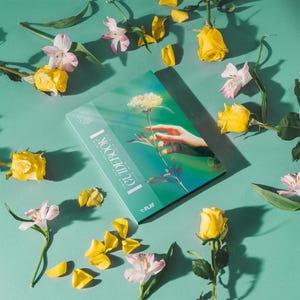 May include: A rectangular guidebook with a teal cover featuring a graphic of a hand reaching towards a yellow flower. The text "GUIDEBOOK I" and "PLAY" are printed on the front. The book is surrounded by scattered yellow roses and pink alstroemeria.