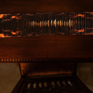 May include: A rectangular, dark-colored object with a fiery, cloud-like pattern on the ends and a central design of vertical lines. The object is set against a dark wooden surface, with a glimpse of a carved wooden chair below.