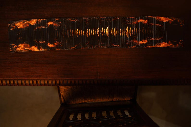 May include: A rectangular, dark-colored object with a fiery, cloud-like pattern on the ends and a central design of vertical lines. The object is set against a dark wooden surface, with a glimpse of a carved wooden chair below.