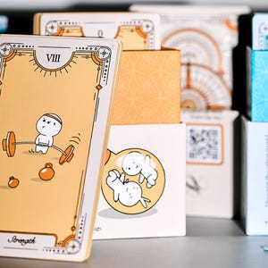 May include: A stack of illustrated cards and boxes. The front card features a cartoon figure lifting weights. The card is tan with a decorative border and the word "Stomach" at the bottom. Other boxes have illustrations of cartoon figures and geometric patterns.