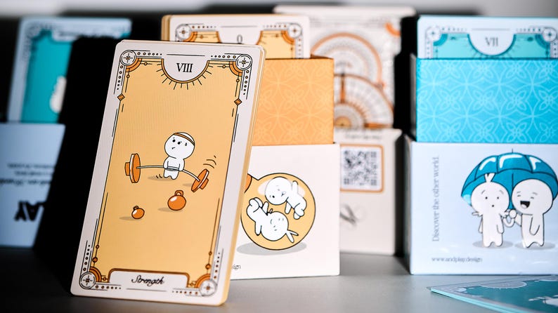 May include: A stack of illustrated cards and boxes. The front card features a cartoon figure lifting weights. The card is tan with a decorative border and the word "Stomach" at the bottom. Other boxes have illustrations of cartoon figures and geometric patterns.