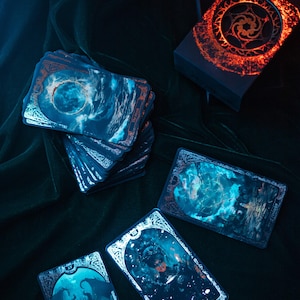 May include: A deck of tarot cards with celestial and mystical imagery. The cards feature blue and teal illustrations with ornate borders and copper-colored accents. A box with a glowing orange design sits nearby.