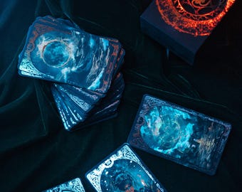 Op de afbeelding: Een tarotkaartspel met hemelse en mystieke beelden. De kaarten tonen blauwe en turquoise illustraties met sierlijke randen en koperkleurige accenten. Een doos met een gloeiend oranje ontwerp is ook zichtbaar.