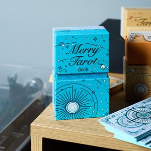 Może przedstawiać: Zestaw talii i pudełek do kart tarota. Przednie pudełko jest turkusowe z napisem "Merry Tarot deck" w białym kolorze. Inne pudełka są beżowe i złote z niebiańskimi wzorami. Talia kart jest rozłożona.