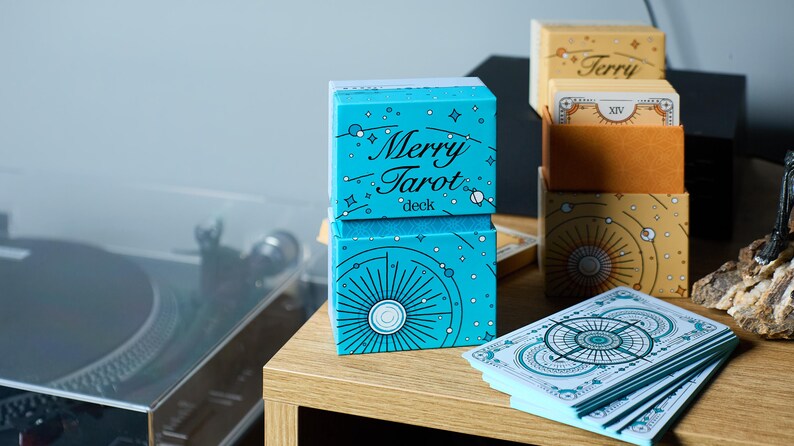 Może przedstawiać: Zestaw talii i pudełek do kart tarota. Przednie pudełko jest turkusowe z napisem "Merry Tarot deck" w białym kolorze. Inne pudełka są beżowe i złote z niebiańskimi wzorami. Talia kart jest rozłożona.