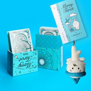 Może przedstawiać: Jasnoniebieski zestaw kart Merry Tarot z przewodnikiem i pluszowym urokiem. Pudełko i przewodnik zawierają ilustracje kr&oacute;lika, ptaka i niebiańskich wzor&oacute;w. Karty tarota mają szczeg&oacute;łowy wz&oacute;r r&oacute;ży wiatr&oacute;w.