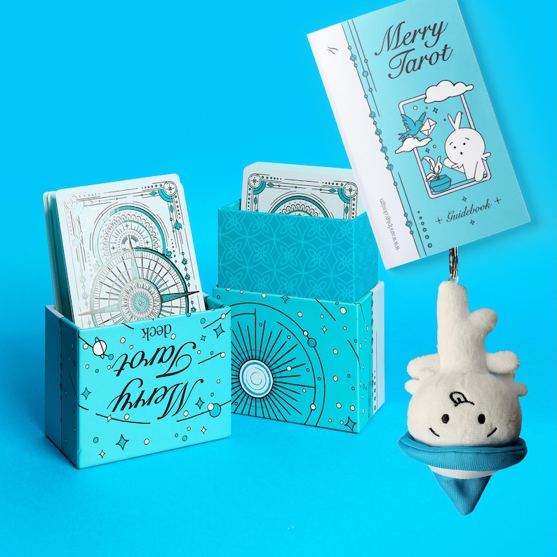 Może przedstawiać: Jasnoniebieski zestaw kart Merry Tarot z przewodnikiem i pluszowym urokiem. Pudełko i przewodnik zawierają ilustracje kr&oacute;lika, ptaka i niebiańskich wzor&oacute;w. Karty tarota mają szczeg&oacute;łowy wz&oacute;r r&oacute;ży wiatr&oacute;w.