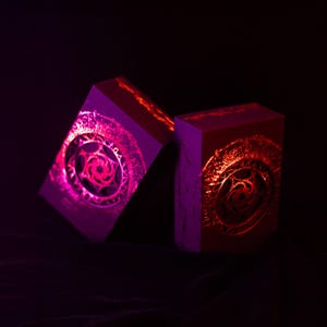 May include: Two rectangular boxes with intricate circular designs, illuminated with vibrant pink and red light against a dark background. The designs appear to be etched or embossed, creating a textured effect.