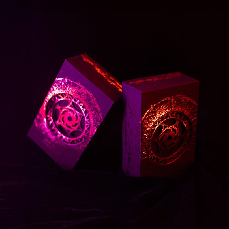 May include: Two rectangular boxes with intricate circular designs, illuminated with vibrant pink and red light against a dark background. The designs appear to be etched or embossed, creating a textured effect.