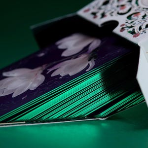 May include: A deck of playing cards partially removed from a white tuck box with intricate cutouts and gold foil accents. The cards feature a dark purple back design with white floral illustrations and metallic emerald green edges.