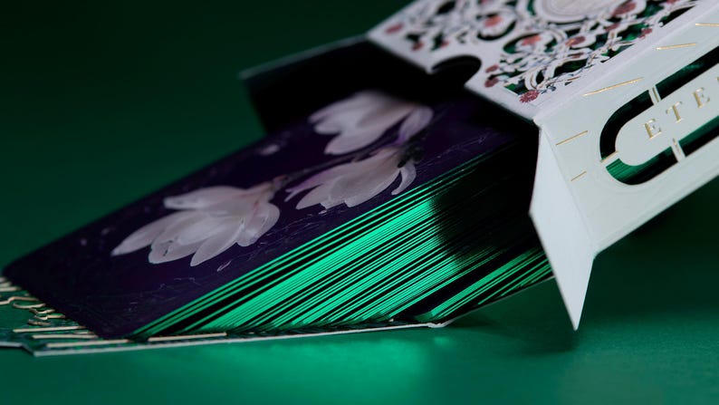 May include: A deck of playing cards partially removed from a white tuck box with intricate cutouts and gold foil accents. The cards feature a dark purple back design with white floral illustrations and metallic emerald green edges.