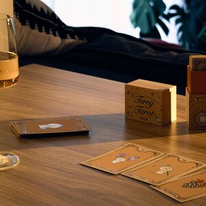 May include: A wine glass filled with a golden liquid sits on a wooden table next to a deck of Terry Tarot cards. The deck is in a wooden box with the words "Terry Tarot" printed on it. Several tarot cards are spread out.