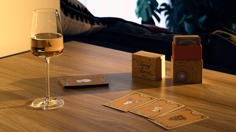 May include: A wine glass filled with a golden liquid sits on a wooden table next to a deck of Terry Tarot cards. The deck is in a wooden box with the words "Terry Tarot" printed on it. Several tarot cards are spread out.