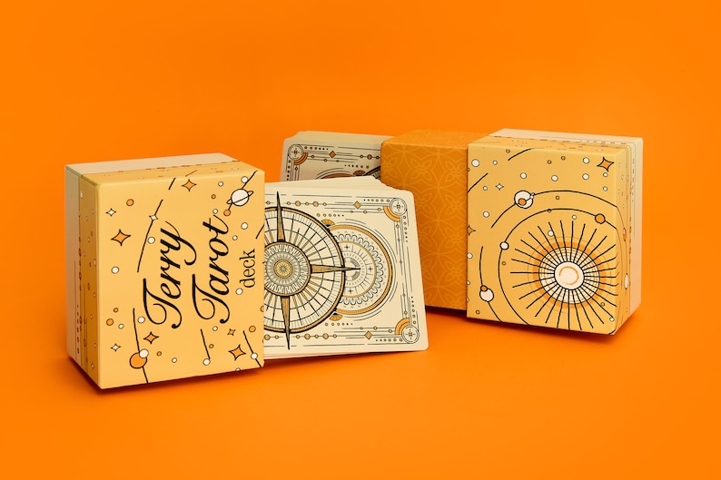 May include: A tarot deck with a light yellow box featuring the text "Terry Tarot deck" and celestial illustrations. The deck includes cards with compass and celestial designs. The background is a solid orange.