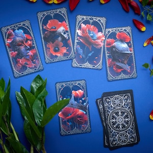 May include: A set of rectangular tarot cards featuring dark, ornate borders and detailed illustrations of red poppies, a skull, a knight's helmet, and a crown. One card shows the back design with a circular, intricate silver pattern on black.