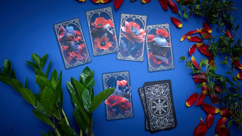 May include: A set of rectangular tarot cards featuring dark, ornate borders and detailed illustrations of red poppies, a skull, a knight's helmet, and a crown. One card shows the back design with a circular, intricate silver pattern on black.