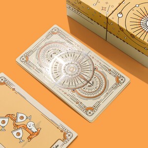 May include: A tarot card deck with a celestial design, featuring cream-colored cards with intricate illustrations in orange and brown. The deck is displayed with a matching box on an orange surface. One card shows three cups and a ghost.